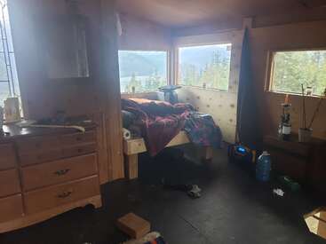 Una acogedora habitación tipo cabaña de madera con grandes ventanas que ofrecen vistas a la montaña y al bosque, una cama deshecha, una cómoda, objetos esparcidos por el suelo y luz natural.