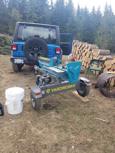 Hay un Jeep azul aparcado frente a una pila de leña en un claro del bosque, junto a una cortadora de troncos Yardworks, un cubo blanco, una silla y un bidón de aceite.