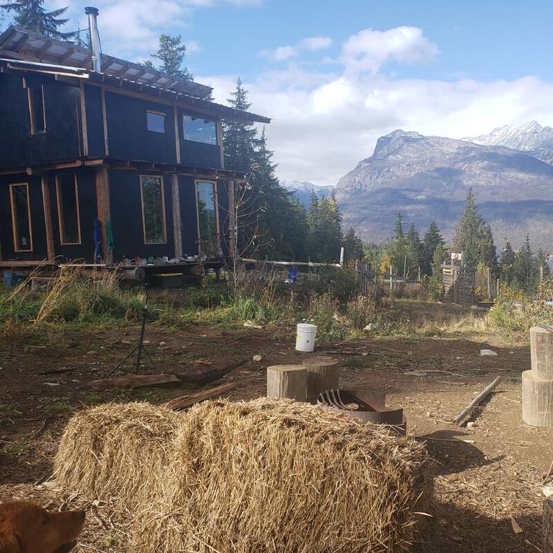 Uma cabana rústica fica em meio a uma clareira na floresta, com fardos de feno em primeiro plano e montanhas majestosas à distância, sob um céu parcialmente nublado.