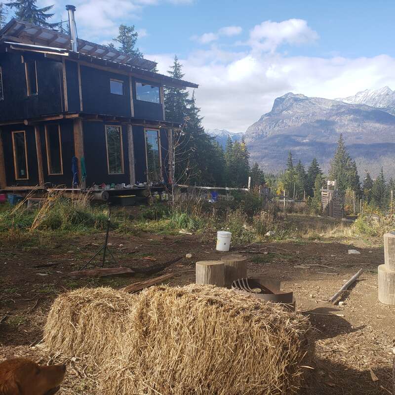 Une cabane rustique se trouve au milieu d'une clairière, avec des bottes de foin au premier plan et des montagnes majestueuses au loin, sous un ciel partiellement nuageux.