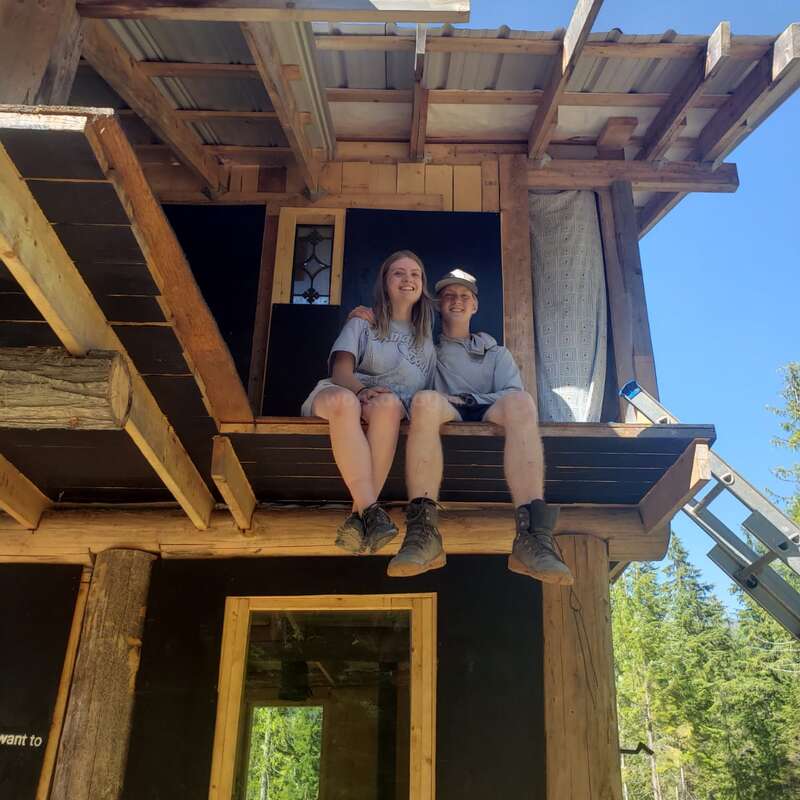Duas pessoas sentadas juntas na varanda inacabada de uma cabana de madeira, sorrindo sob um céu azul claro, cercadas pela floresta, apreciando o progresso da construção.