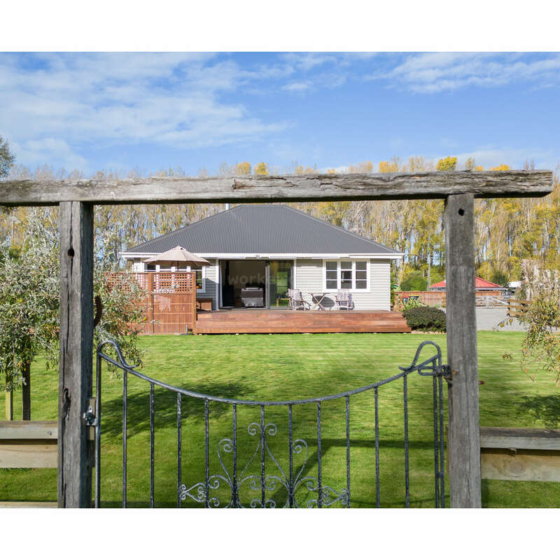 Hinter einem hölzernen Gartentor befindet sich ein charmantes Haus mit einer großen Rasenfläche, einer einladenden Terrasse, Sitzgelegenheiten im Freien und umgeben von Bäumen unter einem strahlend blauen Himmel.
