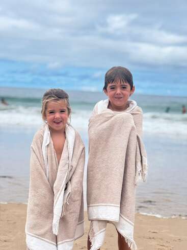 Duas crianças sorridentes estão em uma praia de areia, enroladas em toalhas bege. As ondas do mar e o céu nublado proporcionam um cenário sereno para esse momento alegre.