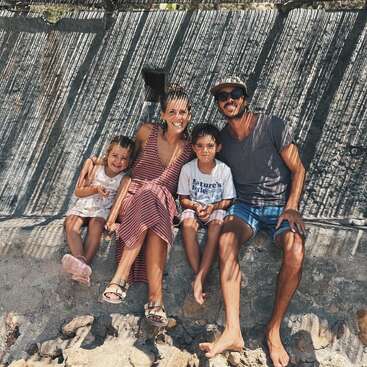 Uma família sorridente de quatro pessoas senta-se ao ar livre em um muro de pedra, desfrutando de um dia ensolarado. As sombras de uma cerca criam padrões listrados sobre eles. Momento feliz.