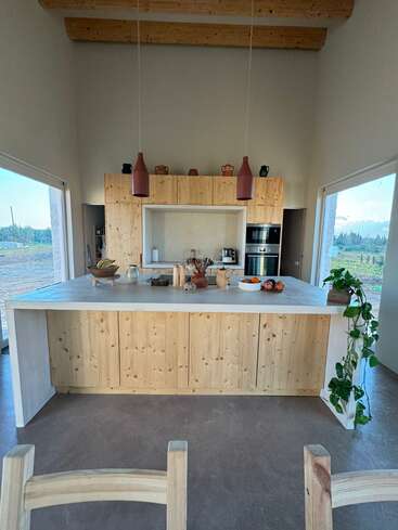Esta imagem mostra uma cozinha rústica moderna com armários de madeira clara, uma ilha grande, decoração minimalista, luzes pendentes, janelas grandes e vistas panorâmicas para o exterior. Aconchegante e convidativo.