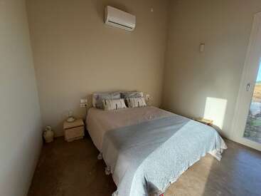 Esta imagem mostra um quarto minimalista com uma cama de casal, roupas de cama leves, duas pequenas mesinhas de cabeceira, ar-condicionado de parede e luz do sol entrando por uma grande janela.