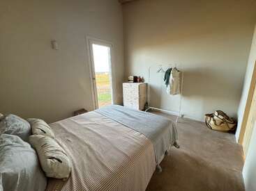 Um quarto minimalista com paredes neutras, uma cama bem arrumada, uma pequena cômoda de madeira, um cabideiro simples, uma janela grande e uma cesta de tecido no chão.
