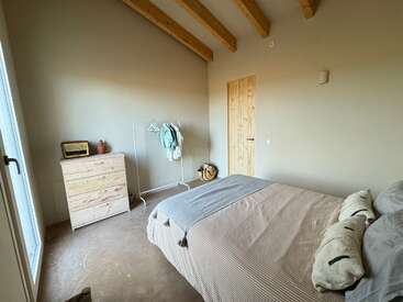Um quarto aconchegante e minimalista com uma cama de madeira, luz natural, tons neutros, vigas de madeira expostas, uma cômoda, um cabideiro e uma decoração simples, criando uma atmosfera calma e convidativa.