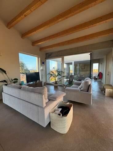 Esta sala de estar aconchegante apresenta sofás neutros, vigas de madeira, janelas grandes, uma TV, plantas de interior, uma cesta de tecido e um design moderno, convidativo e iluminado pelo sol em um espaço aberto.