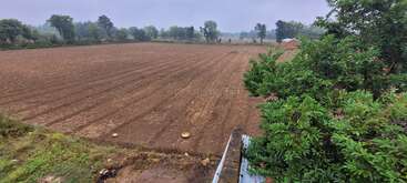 This image shows a freshly plowed agricultural field bordered by green trees, with a small shed in the distance under a cloudy sky on a rural day.