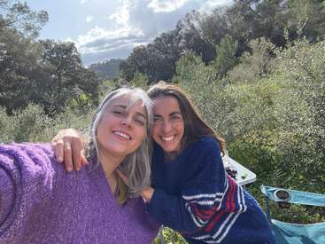Duas mulheres, sorridentes e abraçadas, aproveitam um dia ensolarado ao ar livre. Elas vestem blusas aconchegantes, rodeadas por uma vegetação exuberante e árvores, com o céu azul e as nuvens acima.