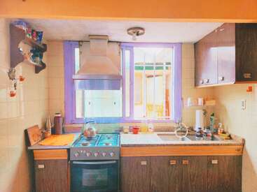 This kitchen features a stove, oven, and range hood, a window with purple trim, wooden cabinets, a sink, countertop appliances, and various kitchen utensils and items.
