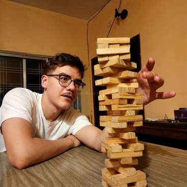 A young man with glasses, wearing a white shirt, carefully plays Jenga. He focuses intensely, reaching to remove a block from the unstable wooden tower.