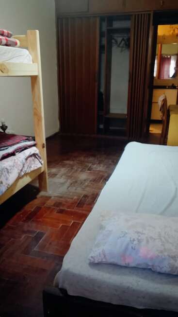 Esta imagem mostra um quarto simples com piso de madeira, uma cama de solteiro, beliches, um armário aberto e um espelho refletindo as cortinas e uma luz.