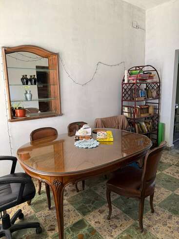 Esta imagem mostra um cômodo aconchegante com uma mesa de jantar vintage de madeira, cadeiras de estilos diferentes, um espelho, uma estante com livros e jogos e luzes de corda na parede.