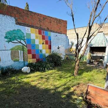 A sunny backyard with green grass, a tree, a colorful mural on the wall, plants, an outdoor grill, and a white tipped pot lying sideways.