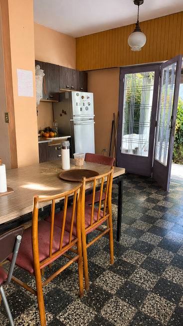 Uma cozinha aconchegante com paredes laranja, geladeira vintage, cadeiras e mesa de madeira, piso de azulejos xadrez e portas de vidro abertas, que deixam entrar a luz natural e oferecem vista para o jardim.