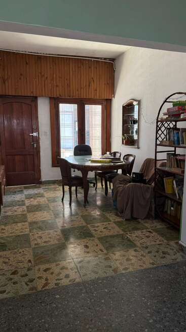 Um quarto aconchegante com uma mesa de madeira, quatro cadeiras, uma estante cheia de livros, um espelho na parede, ladrilhos estampados e a luz do sol entrando pelas janelas.