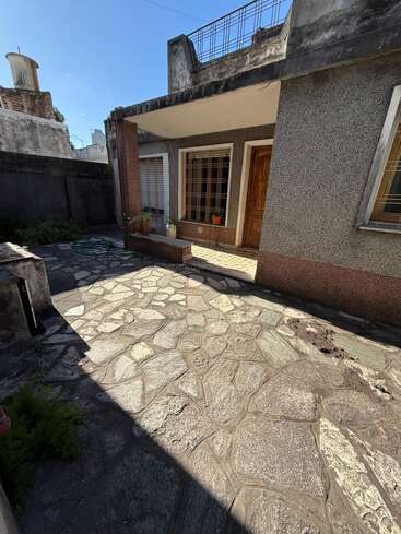 Um pátio ensolarado, com piso de pedra e alguns vasos de plantas, que leva a uma casa com janelas amplas, uma porta de madeira e paredes externas desgastadas pelo tempo.