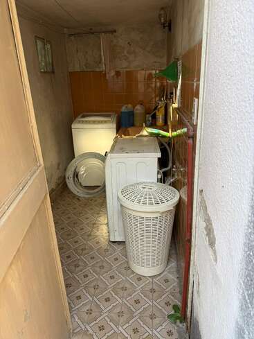 Esta é uma pequena lavanderia com uma máquina de lavar, um cesto de roupa suja, produtos de limpeza, paredes revestidas com azulejos marrons, piso estampado e alguns canos à vista e paredes antigas.