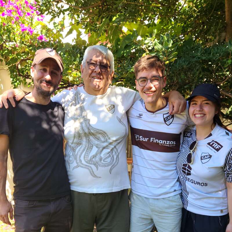 Four people stand closely together outdoors, smiling warmly. They are wearing casual clothes and three have matching football shirts. Sun filters through leaves, creating a cheerful, relaxed atmosphere.