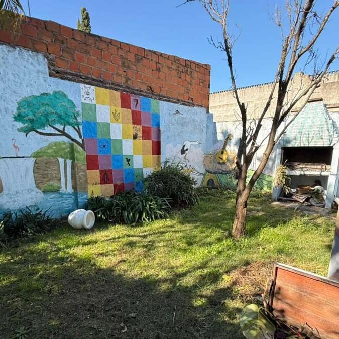Um quintal ensolarado com grama verde, uma árvore, um mural colorido na parede, plantas, uma churrasqueira ao ar livre e um vaso com a borda branca deitado de lado.
