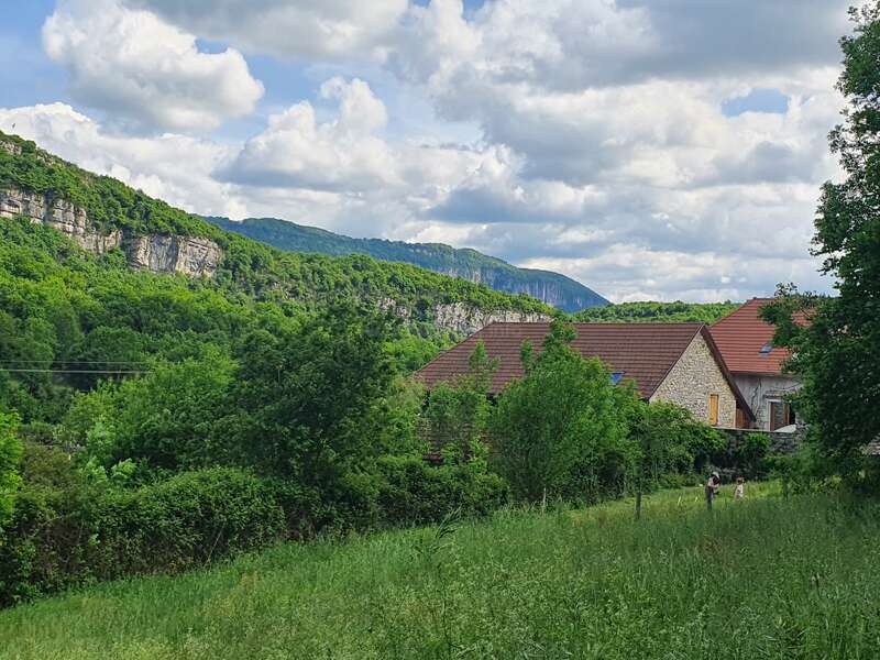 Das Bild zeigt eine ruhige Landschaft mit einem Haus mit braunem Dach, eingebettet in üppiges Grün und hoch aufragende Bäume, mit einer majestätischen Bergkette im Hintergrund.