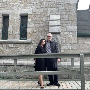 Ein dunkel gekleidetes Paar steht gemeinsam auf einem Holzsteg vor einem Steingebäude mit schmalen Glasfenstern. Bewölktes Licht.