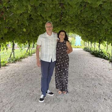 Ein Mann und eine Frau stehen zusammen unter einem üppigen grünen Bogen aus Reben, lächeln und genießen einen friedlichen, sonnigen Tag inmitten von Natur und Grün.