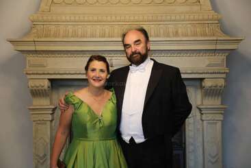 Une femme en robe verte et un homme en smoking se tiennent côte à côte, souriants, devant une cheminée en pierre richement décorée, posant pour une photo.