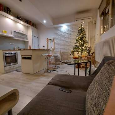 A cozy living space features a modern kitchen, decorated Christmas tree with lights, bar stools, sofa, coffee table, and warm lighting, creating a festive atmosphere.