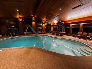 This image shows a cozy indoor pool with a slide, surrounded by warm wooden walls and ceilings, and comfortable seating and tables along the perimeter.