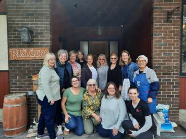 A group of twelve women pose and smile outside a rustic brick building. Some are standing, some kneeling, and they look happy and relaxed together.