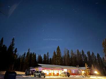 A cozy, brightly lit cabin sits under a clear, starry night sky, surrounded by snow and tall pine trees. Two cars are parked nearby. Peaceful atmosphere.