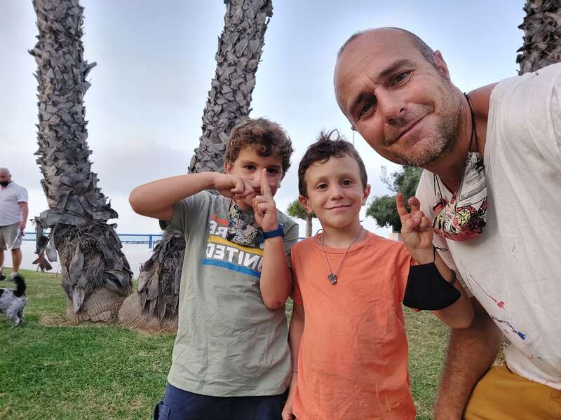 La imagen muestra a un hombre y dos niños posando para un selfie delante de una palmera, con los niños haciendo gestos con las manos.
