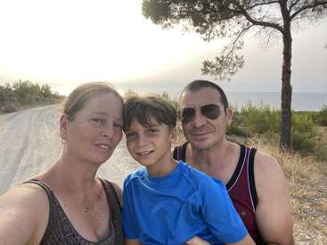 The image depicts a family of three posing for a selfie on a dirt road, with a serene background featuring trees, bushes, and a body of water under an overcast sky.