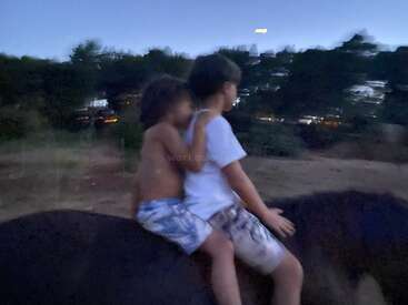 Two children sit on a dark object, with one child\'s arm around the other, set against a blurred background of trees and buildings.