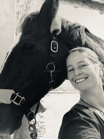 The image is a black-and-white photograph of a woman posing with a horse, both looking at the camera, in front of a stable.