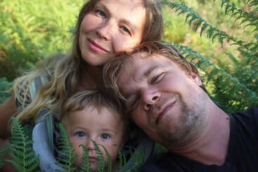 Una sonriente familia de tres miembros -madre, padre e hijo- posan muy juntos entre verdes helechos al aire libre, irradiando calidez, felicidad y amor en la naturaleza.