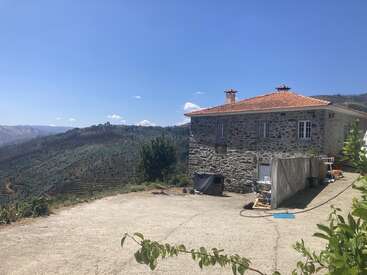 Una rústica casa de piedra con tejado de tejas rojas se asienta en una soleada ladera, con vistas a exuberantes montañas verdes y valles bajo un cielo azul despejado.