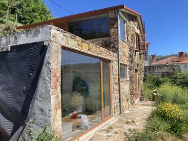 Esta imagen muestra una casa rústica de piedra con grandes ventanales, rodeada de vegetación y flores silvestres bajo un cielo azul despejado, que desprende un apacible ambiente campestre.