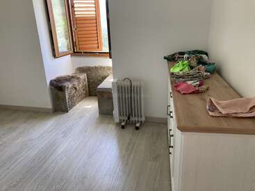 Habitación sencilla y luminosa con una ventana abierta, un banco de piedra, un radiador eléctrico y una cómoda de madera con ropa doblada. El suelo es de madera clara.