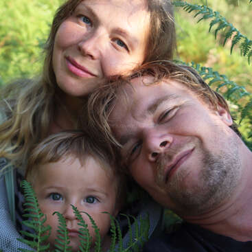 A smiling family of three—mother, father, and child—pose closely together among green ferns outdoors, radiating warmth, happiness, and love in nature.