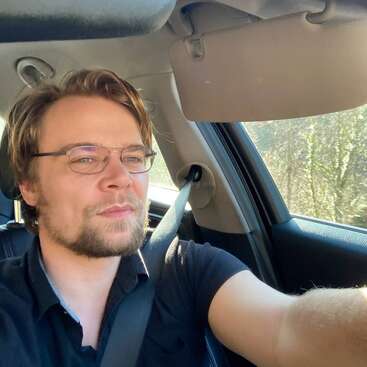 Un hombre con gafas y barba conduce un coche, con el cinturón de seguridad abrochado. La luz del sol entra por la ventanilla e ilumina el interior mientras él mira hacia delante.