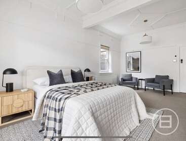 This bedroom features a large bed with checkered blankets, two black lamps, stylish side tables, armchairs, wall art, and neutral colors, creating a cozy modern retreat.