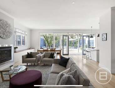This image features a modern, open-plan living area with neutral furniture, a fireplace, large windows, natural light, dining table, kitchen island, and access to a patio.