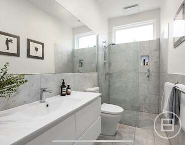 This modern bathroom features a glass-enclosed shower, marble tiles, a sleek white vanity, wall art, large mirror, and natural light from high windows. Minimalist decor.