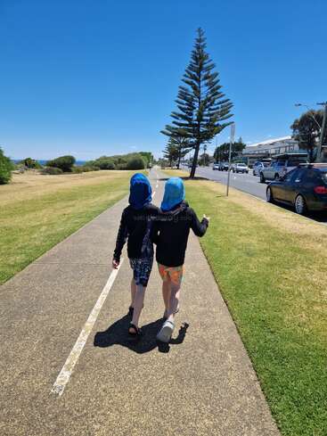 Two children, dressed in sun-protective clothing with blue hats, walk side by side on a sunny path near a road and park, under a clear blue sky.