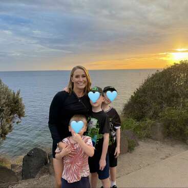 A woman and three children pose happily by the seaside at sunset. The children’s faces are covered with blue heart emojis. Scenic ocean and sky background.