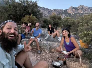 Eine Gruppe von sechs Personen sitzt im Freien, lächelt und genießt eine gemeinsame Mahlzeit. Sie sind von Bäumen und Bergen umgeben, was auf ein zwangloses, fröhliches Picknick hinweist.